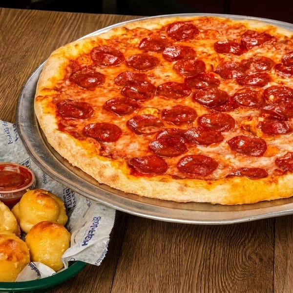 Pepperoni pizza on a metal tray next to a basket of garlic knots and marinara sauce.